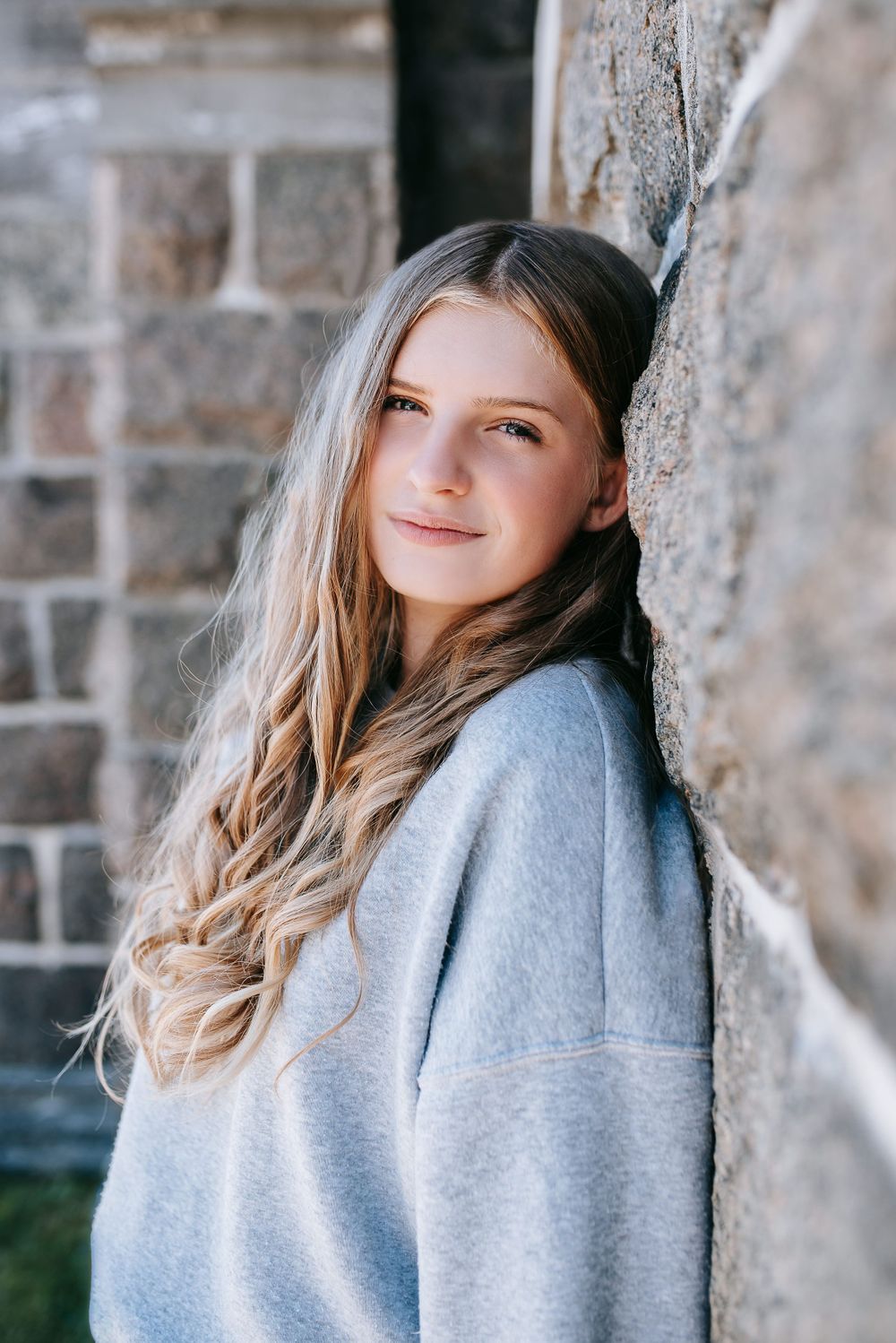 Portrait of person with long blonde hair leaning against brick wall wearing grey sweatshirt.