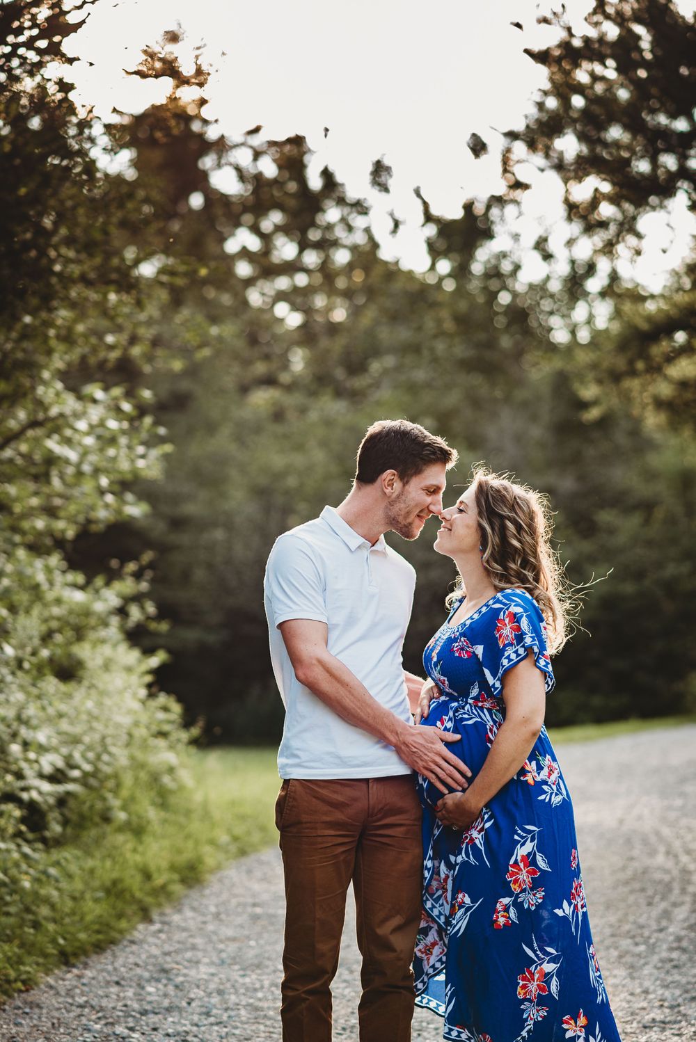 Romantic couple in blue floral dress and brown pants embrace on a sunlit path in nature.