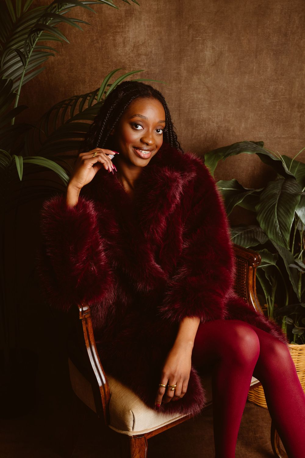 A fashion photoshoot featuring a burgundy faux fur jacket and matching pants in a moody, plant-filled setting.