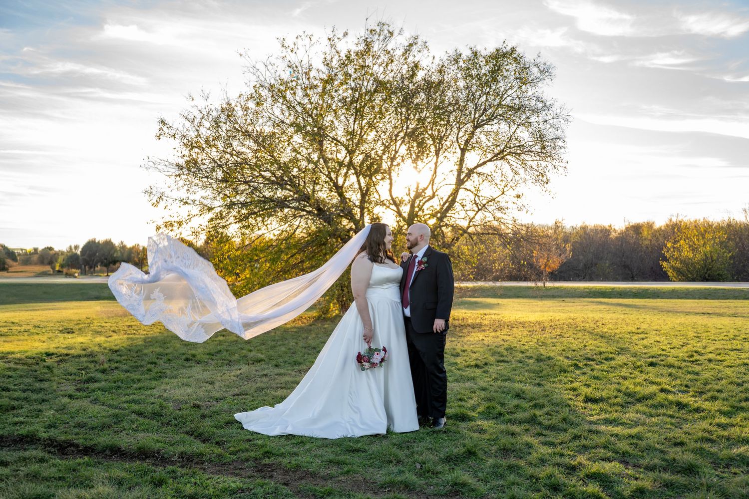 Wedding couple posing in golden sunset light across open field with flowing veil.