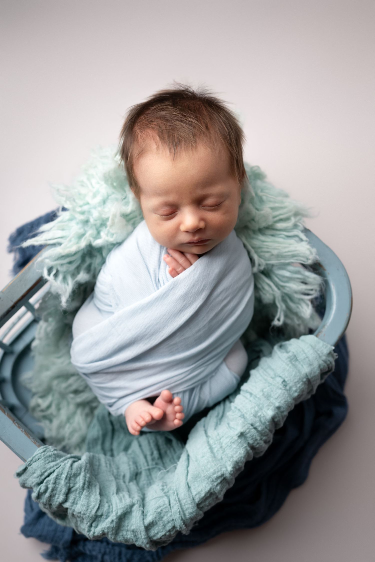 Sleeping newborn swaddled in gray fabric nestled on a teal tulle fabric backdrop in multiple artistic poses.