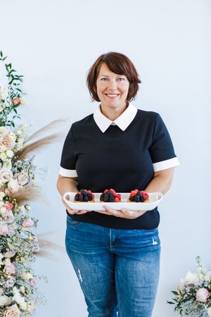 Person in black and white collared shirt holding a plate of colorful food against a floral backdrop.