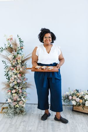 A person in a white sleeveless top and navy shorts sits on a stool next to pink and white floral arrangements.