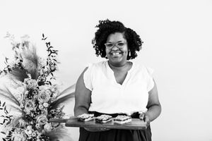 Black and white photo showing someone in a white v-neck top holding a tray of food next to floral arrangements.