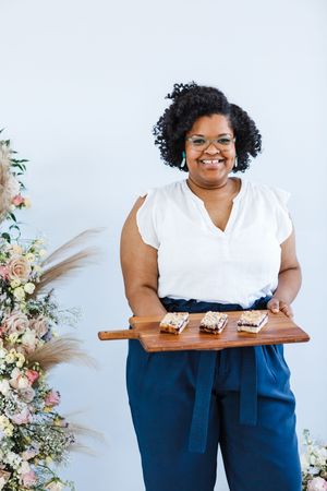 A person wearing a white top and navy pants poses in front of a light backdrop with floral arrangements.