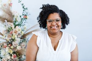 Smiling woman in white dress poses with elegant floral arrangements and pastel decor at professional photo shoot.