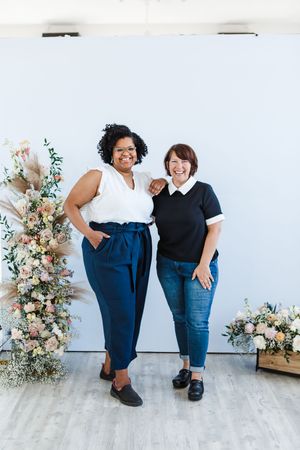 Two friends in black shirts and blue pants pose together against a white wall with floral arrangements.