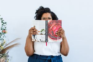 Person holding up a book or magazine while sitting against a white wall with greenery visible in the background.
