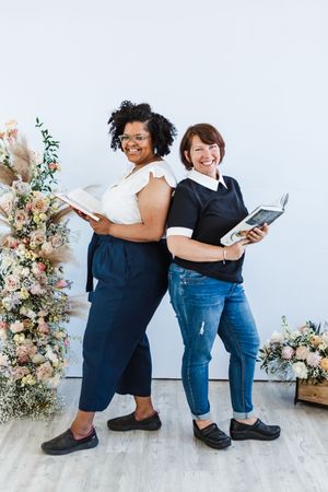 Two people in contrasting outfits stand back to back holding books with floral arrangements in the background.
