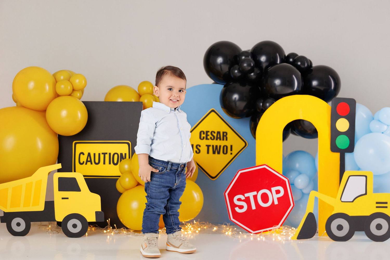 Construction themed birthday party setup with yellow and black balloons, toy trucks, and traffic signs against a gray backdrop.
