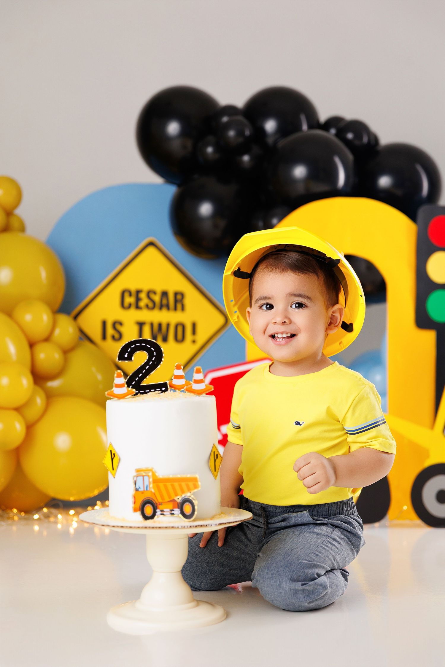A child wearing a yellow construction hat plays with toy trucks surrounded by black and yellow balloons at a birthday party.