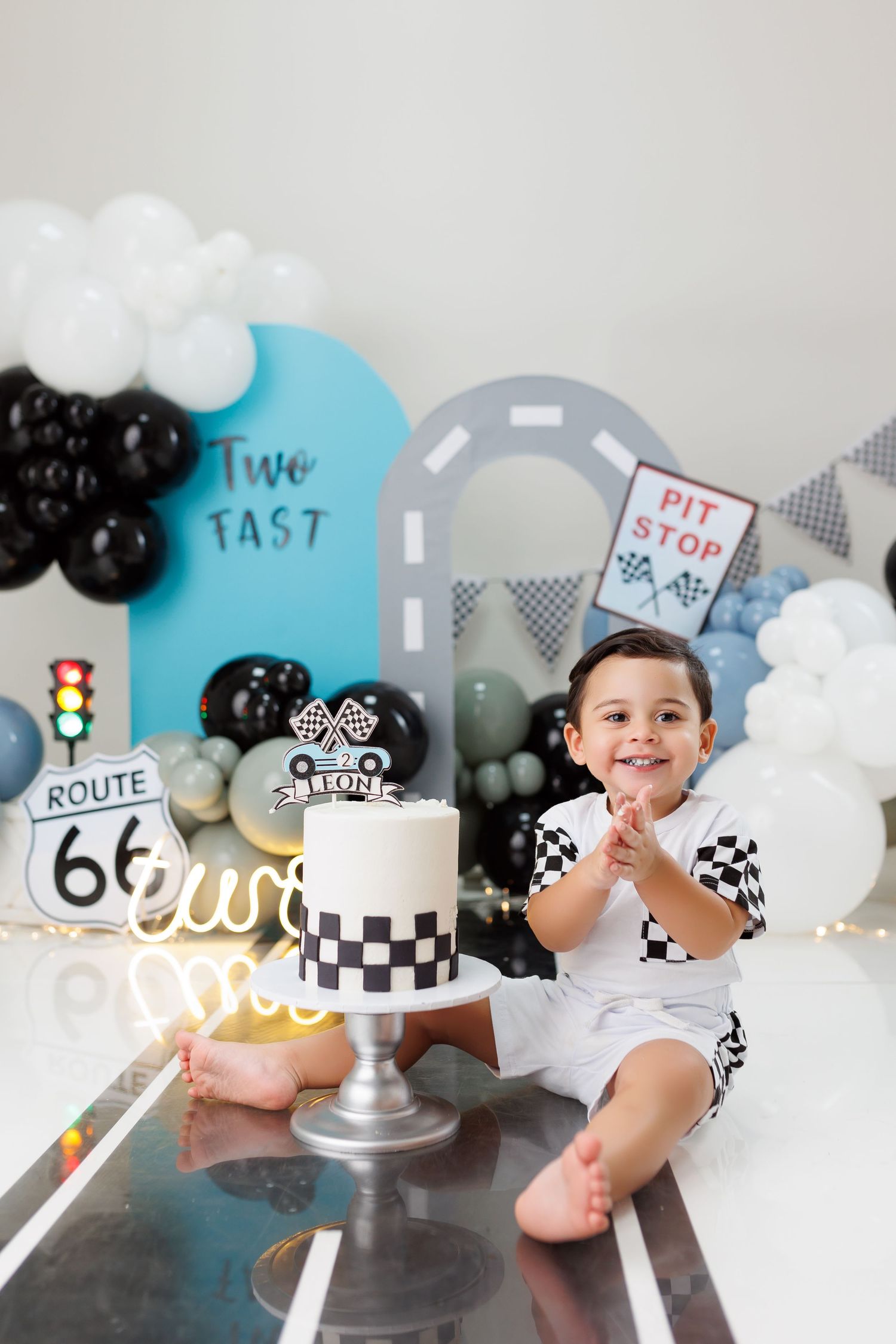 Racing themed birthday party setup with checkered cake, Route 66 sign, and balloon decorations in black, white and blue colors.
