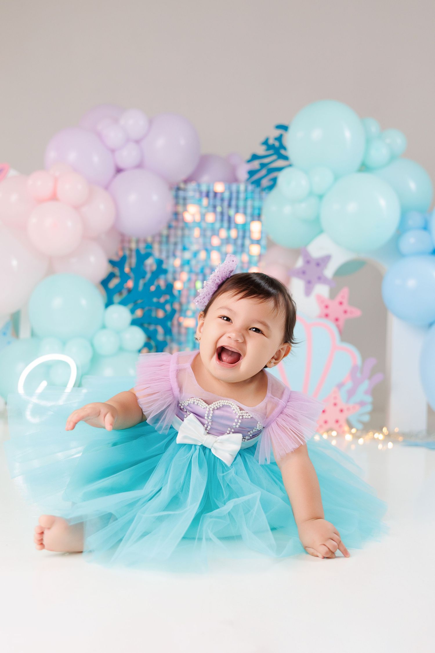 A joyful baby wearing a turquoise tulle dress poses against a pastel balloon arch backdrop with pink and blue decorations.