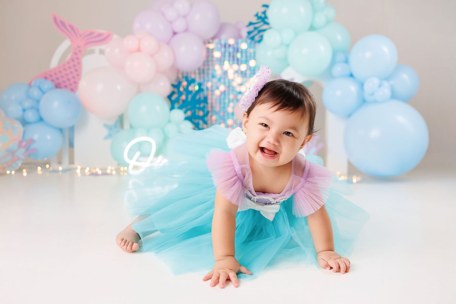 A baby in a teal tutu dress crawls on a white floor with pink and blue balloon decorations in the background.