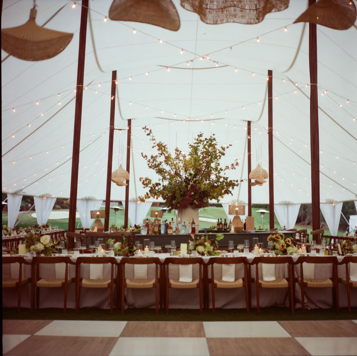 Long banquet tables set with elegant white linens and greenery centerpieces under a white tent for an outdoor wedding reception.