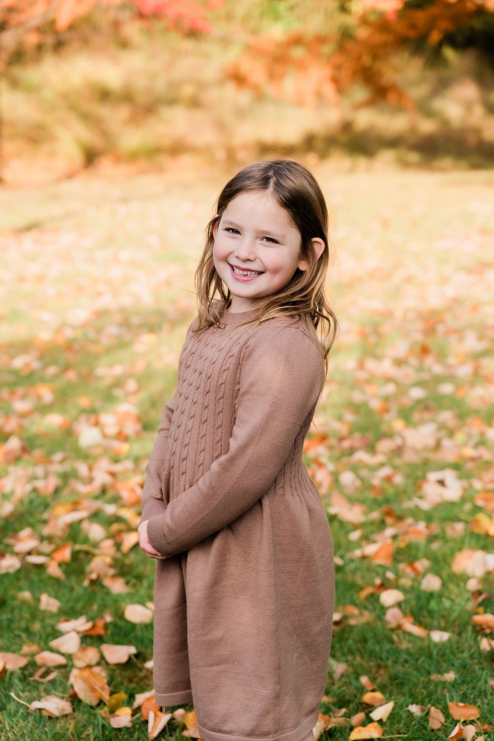 A child wearing a brown sweater plays and smiles in a yard filled with colorful autumn leaves on the ground.