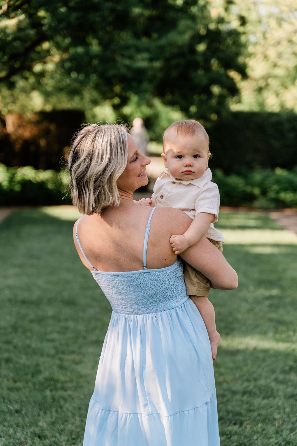 A tender moment captured outdoors as a woman in a light blue sundress holds and interacts with a baby on a sunny day.