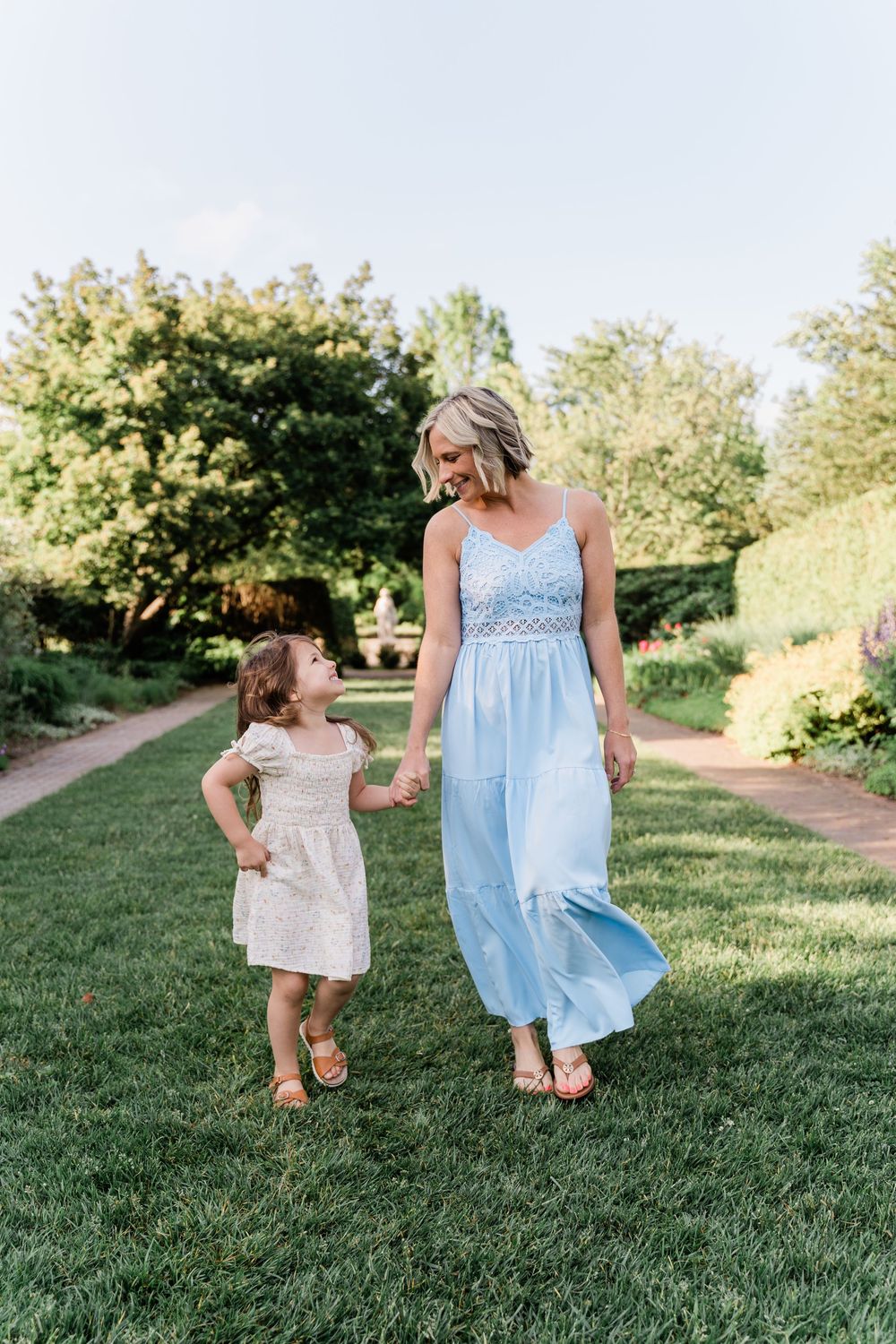 Two people in flowing summer dresses walk and play together on a grassy hillside path on a sunny afternoon.