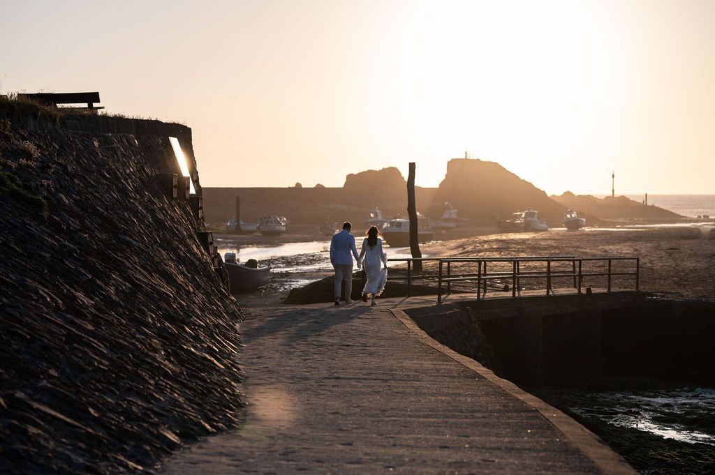 After the wedding ceremony, a couple walk around Bude Beach.  They are heading into the sunset in the distance, with boats in the background