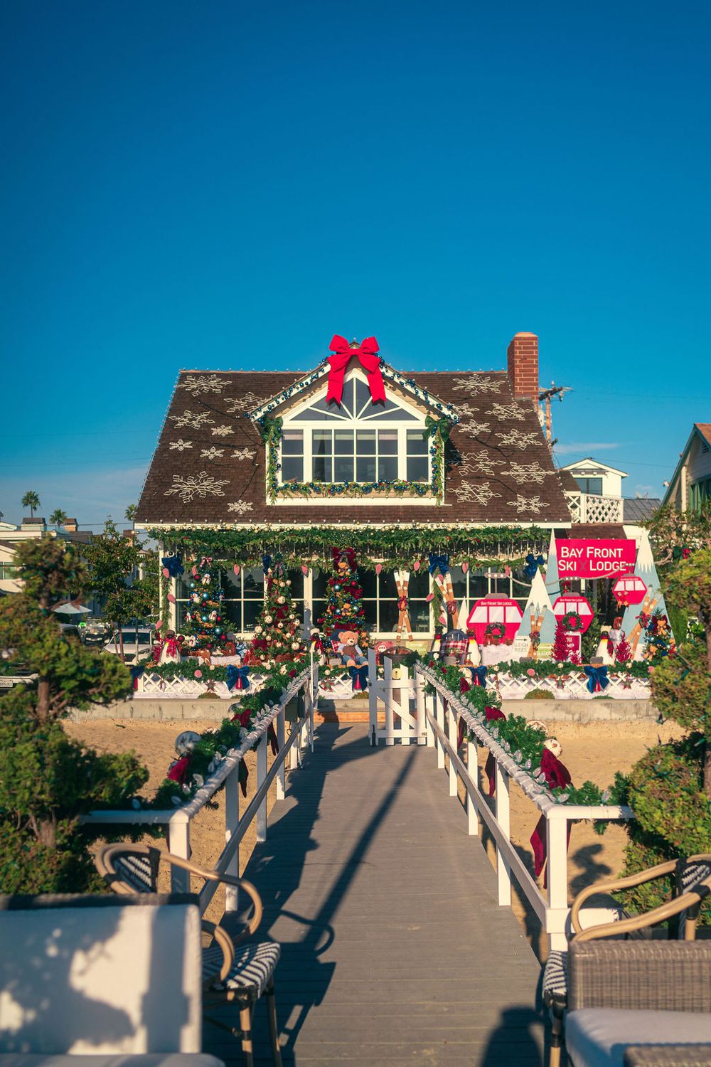 A beachfront walkway decorated with holiday lights and garland leading to a coastal home under bright blue skies.