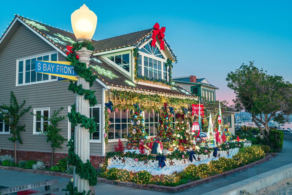 Coastal shopping village with gray buildings decorated in festive Christmas garlands and lights against a bright blue sky.