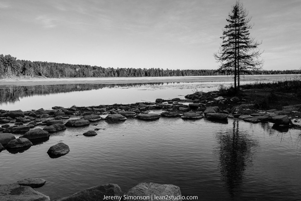 A black and white panoramic landscape of a serene lake with scattered boulders and a lone pine tree silhouetted against the sky.