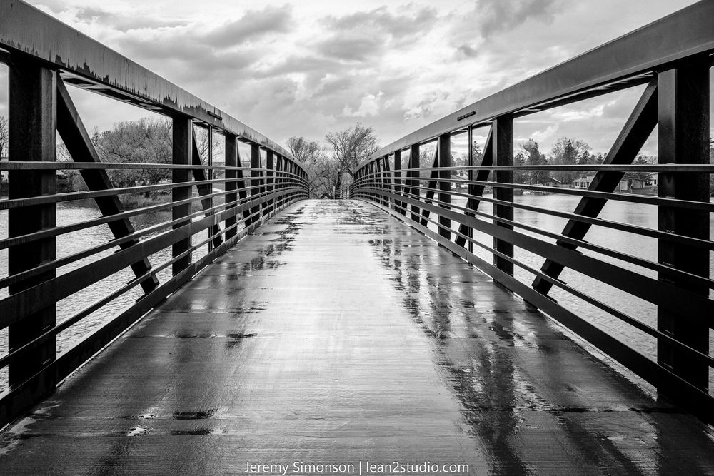 A dramatic black and white photograph of a long wooden boardwalk stretching into the distance under moody storm clouds.