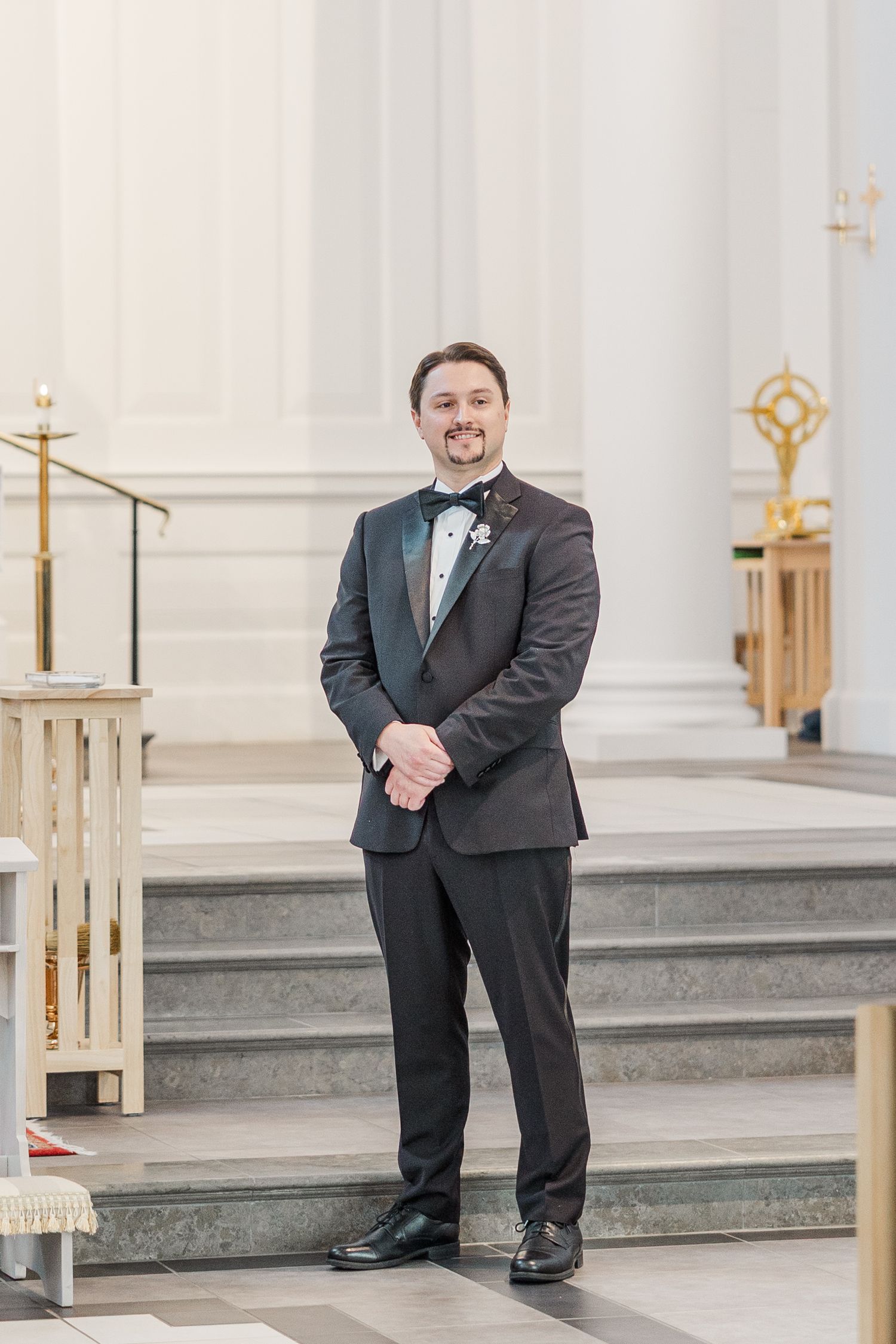 A bride in a white wedding dress walks down the aisle of a bright church with someone in a black tuxedo.