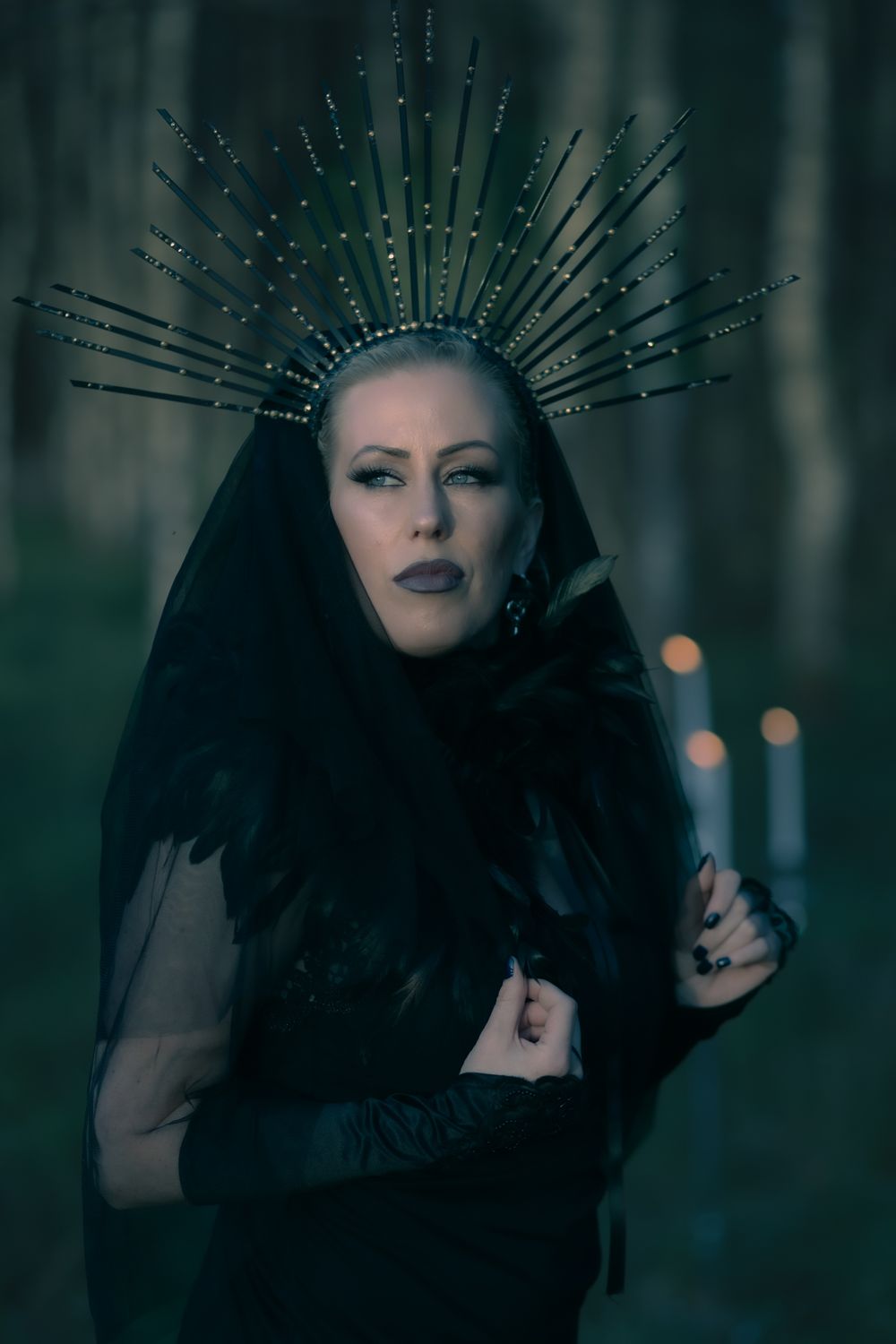 Dark gothic portrait with ornate spiked crown headpiece and black veil in moody forest setting with bokeh lights.