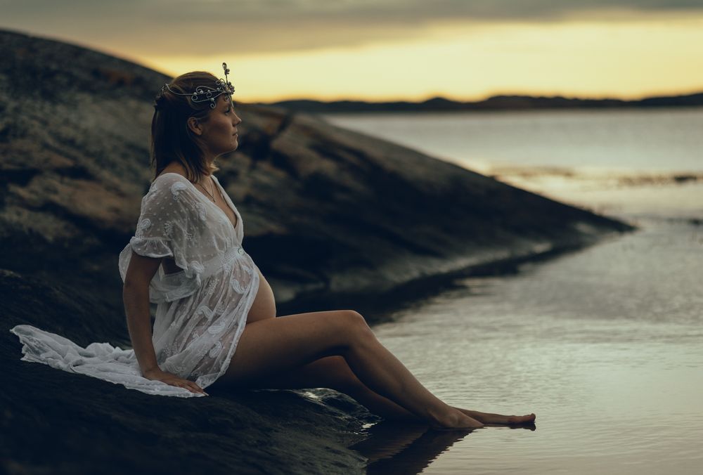 Silhouette of person sitting on rocky coastline at sunset with flowing white fabric creating dreamy atmosphere.