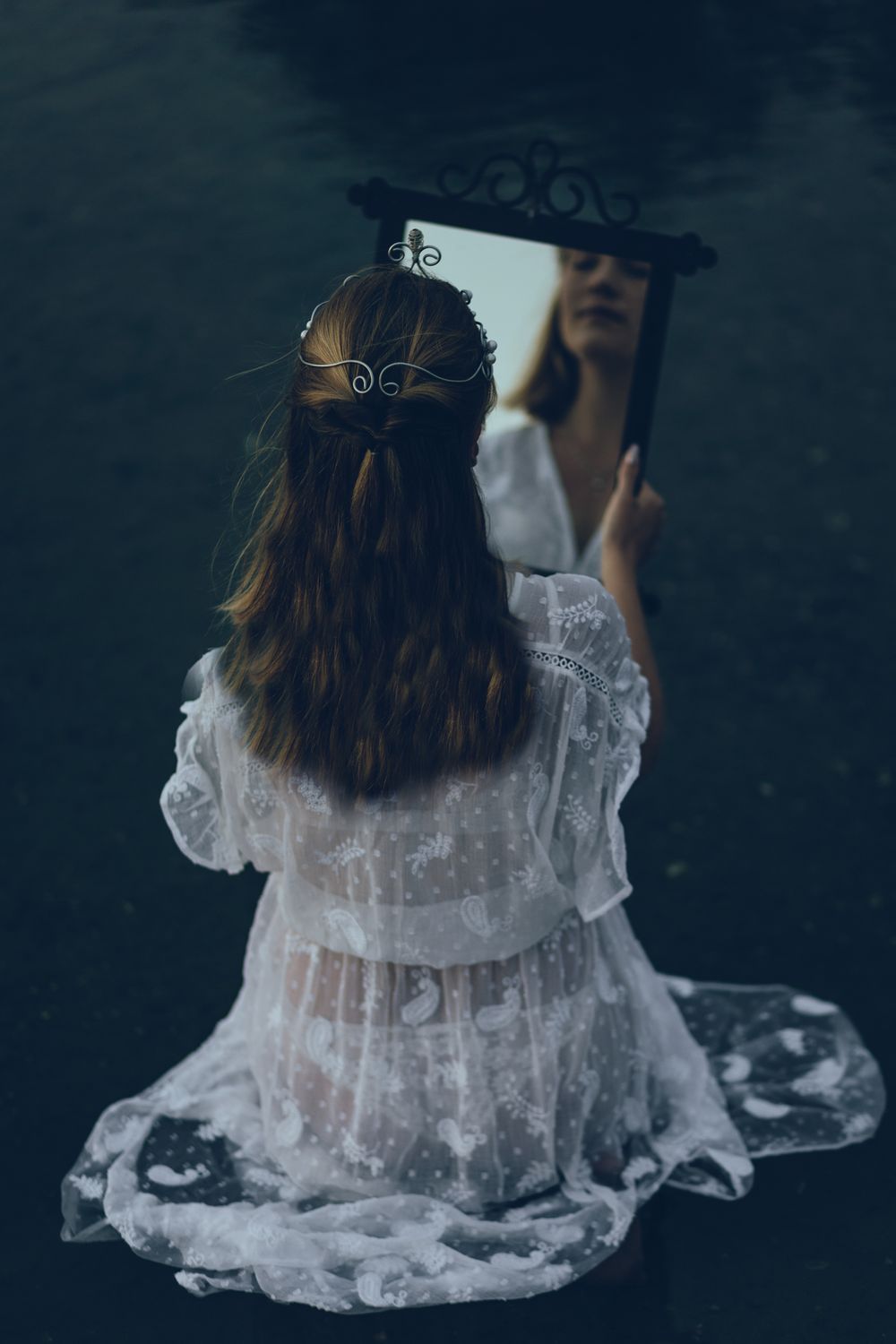 A figure in a white lace dress and crown sits on dark ground creating a moody, ethereal atmosphere.