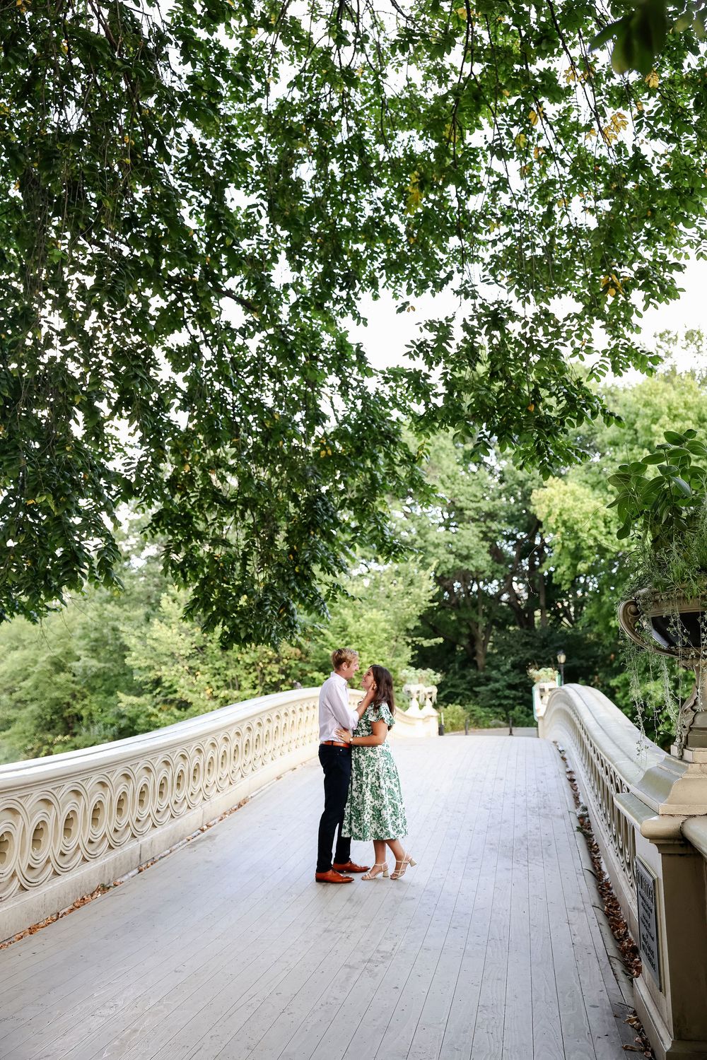 A couple shares a romantic moment on a scenic stone bridge surrounded by lush green trees in a park setting.