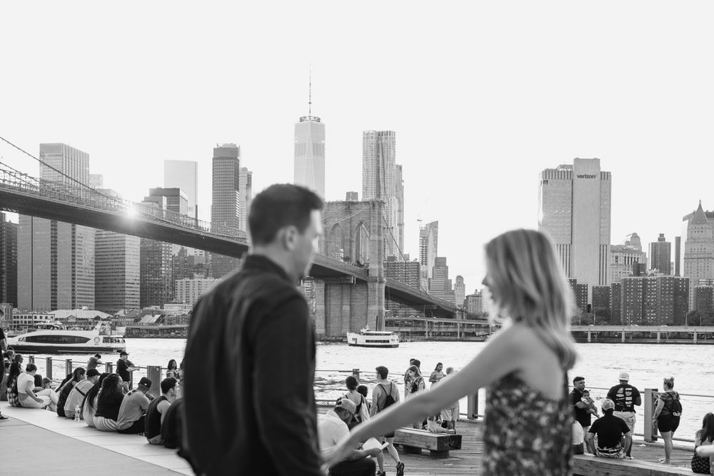 A couple enjoys a scenic moment with the Brooklyn Bridge and Manhattan skyline in the background.