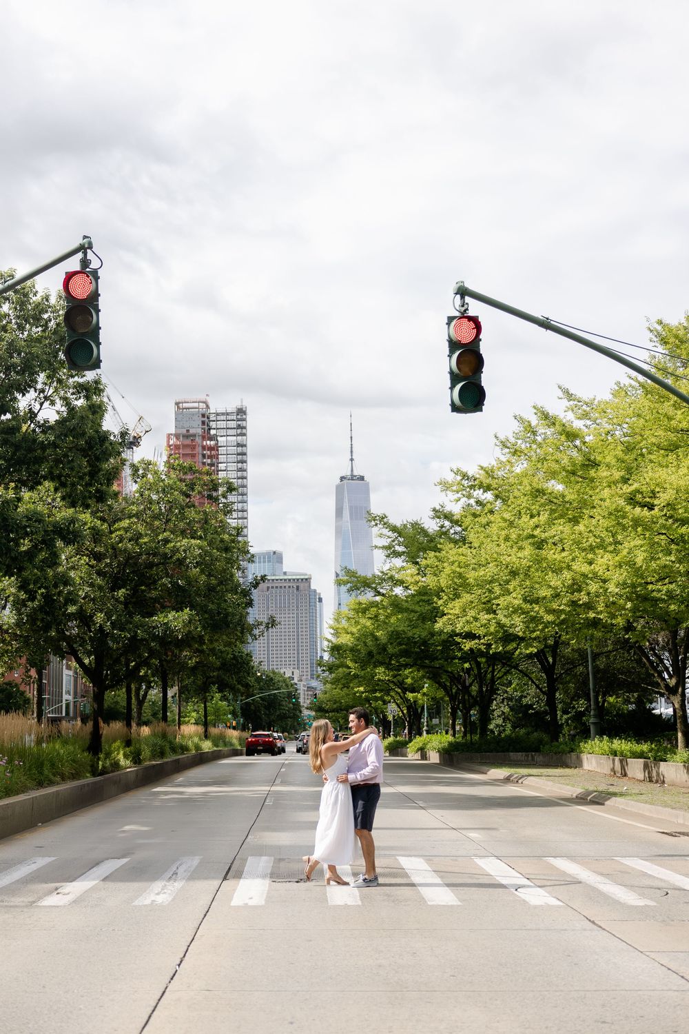 Two people cross a city street with green trees and tall buildings visible in the urban landscape during daytime.