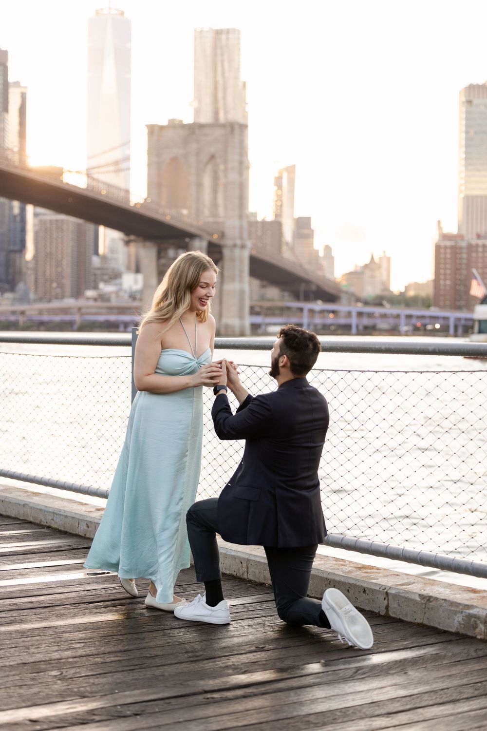 A couple shares a romantic proposal moment on a wooden boardwalk with the Brooklyn Bridge in the background.