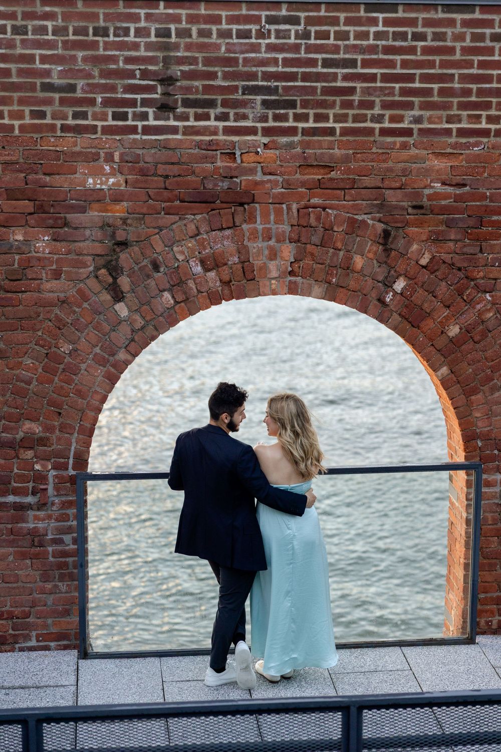 A couple sits together on a bench against a brick wall with arched windows in an urban setting.