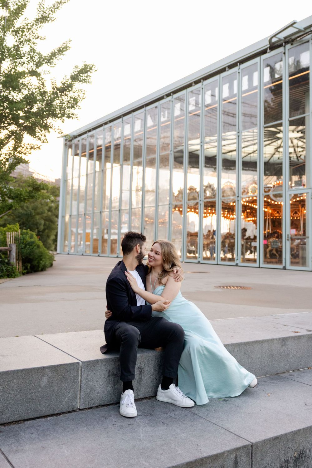 A couple sits together on concrete steps outside a modern glass building at sunset.