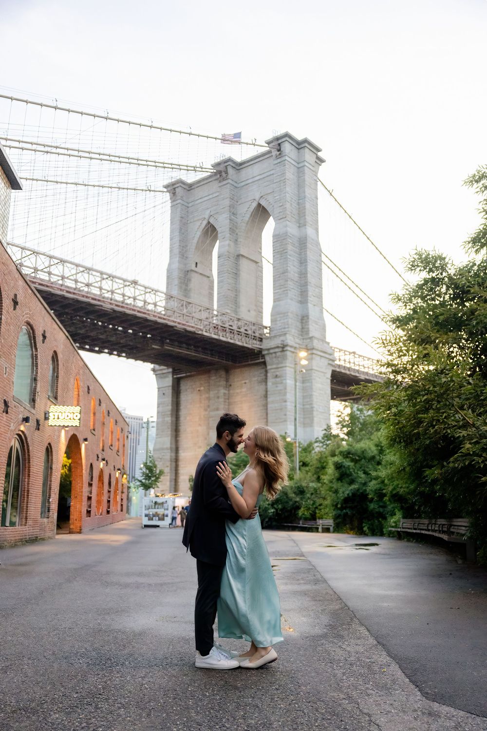 A romantic moment beneath the iconic Brooklyn Bridge with brick buildings and greenery in the background.