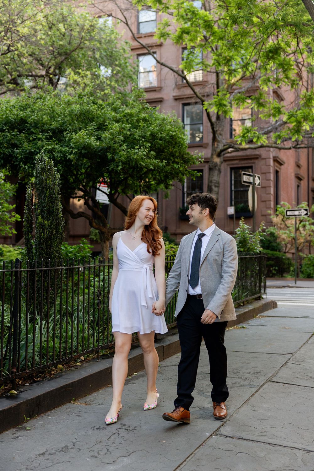 A couple walks together on a tree-lined sidewalk in front of brownstone buildings on a spring day.