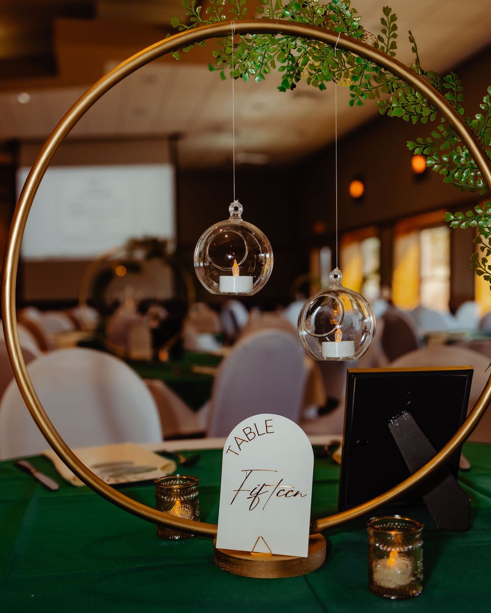Modern wedding table decor features a gold hoop arch with hanging glass orbs and greenery above a table number display.