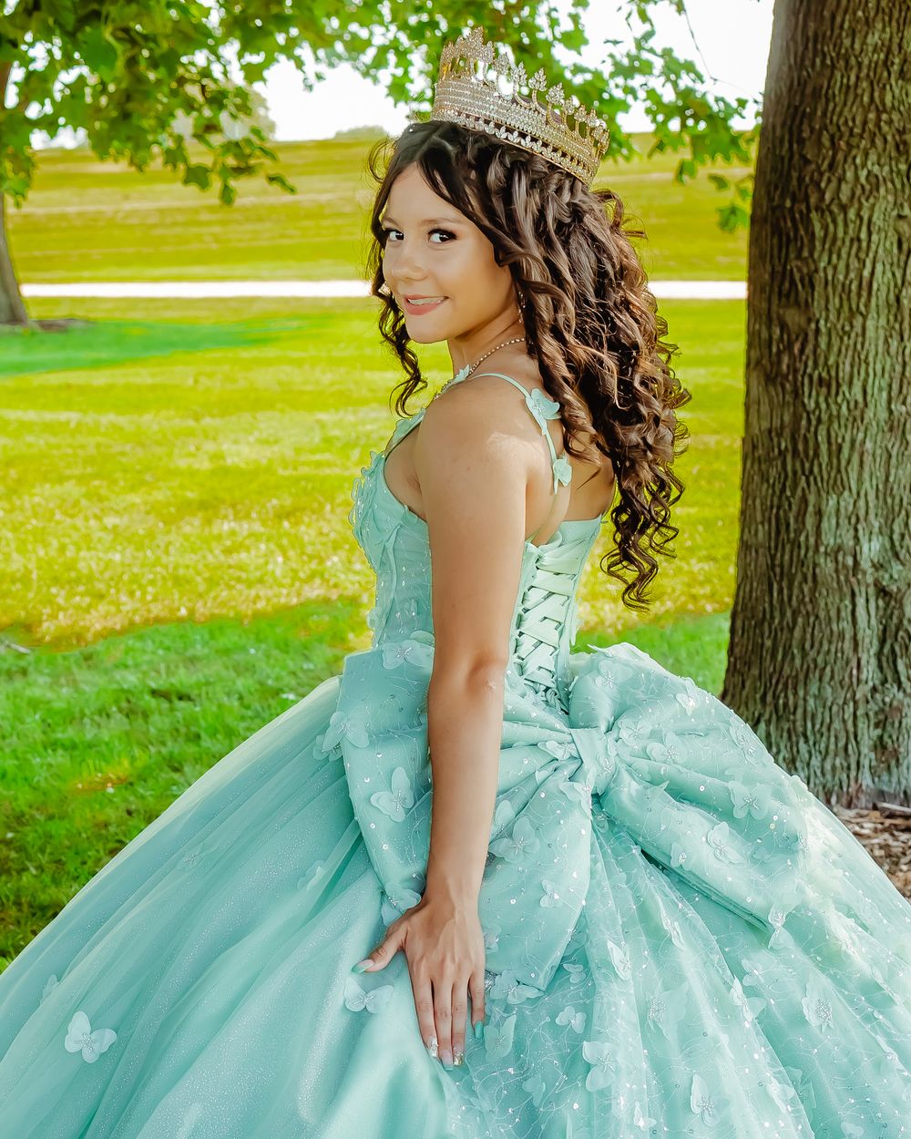 A person in a mint green quinceañera ball gown with curled hair and tiara poses by a tree in a grassy outdoor setting.