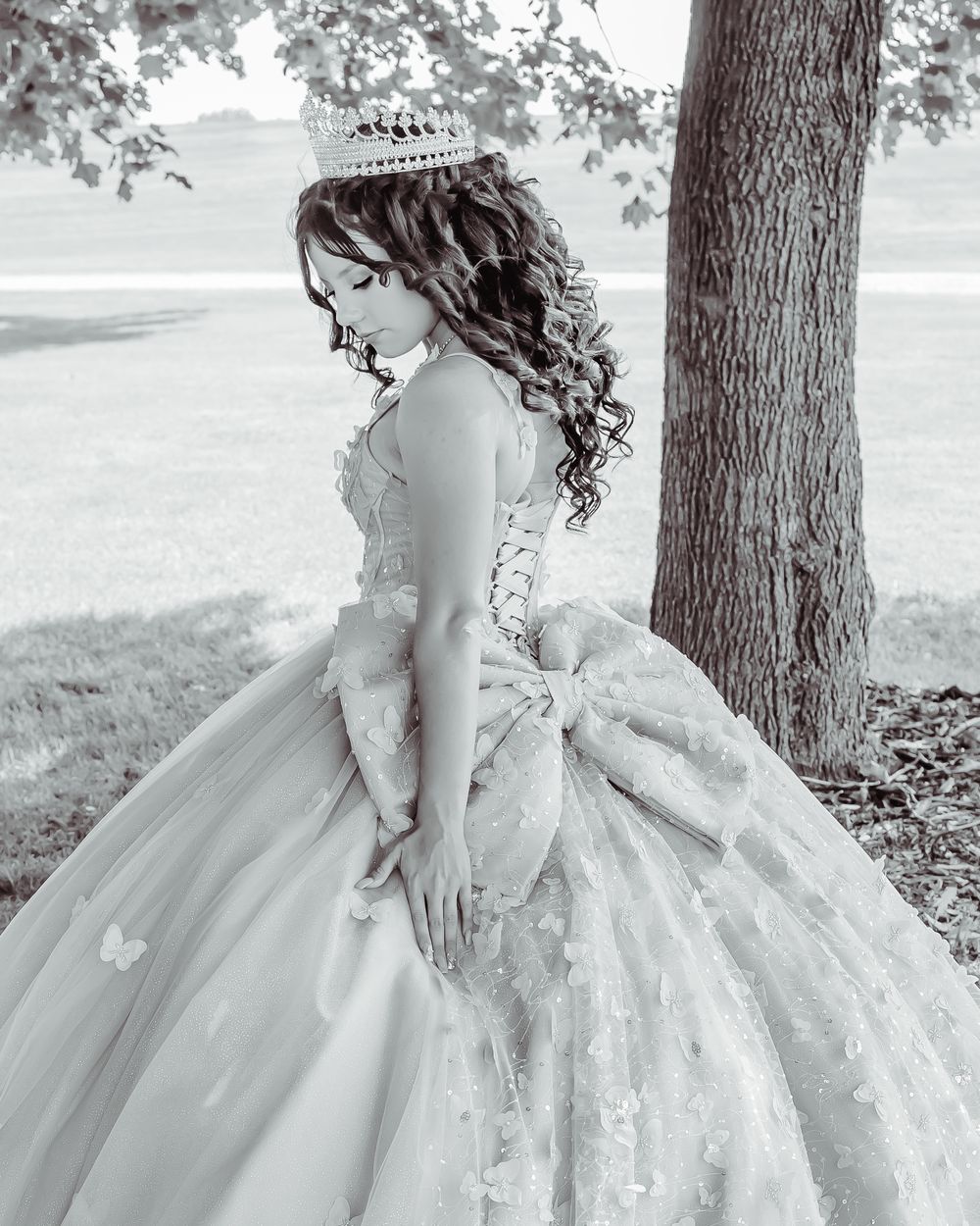 A side profile view of someone in an elegant white ball gown and tiara standing beside a tree in black and white.