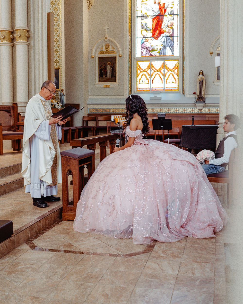 A quinceañera celebration in a Catholic church with a young woman in a pink ballgown kneeling before a priest at the altar.