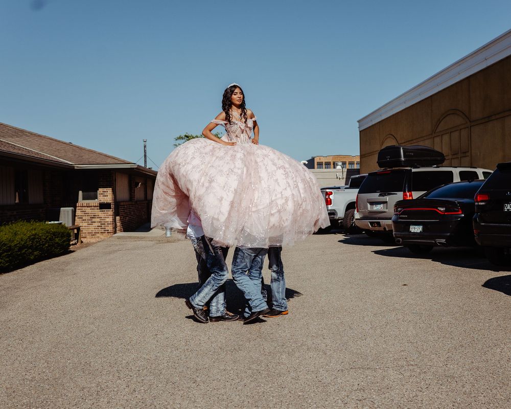 A person in a flowing pink quinceañera dress is lifted by helpers in a parking lot on a sunny day.