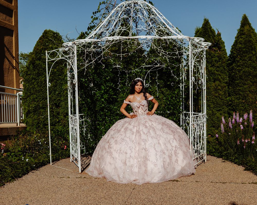 A stunning blush pink quinceañera ball gown displayed under an ornate white metal garden gazebo with twinkling lights.