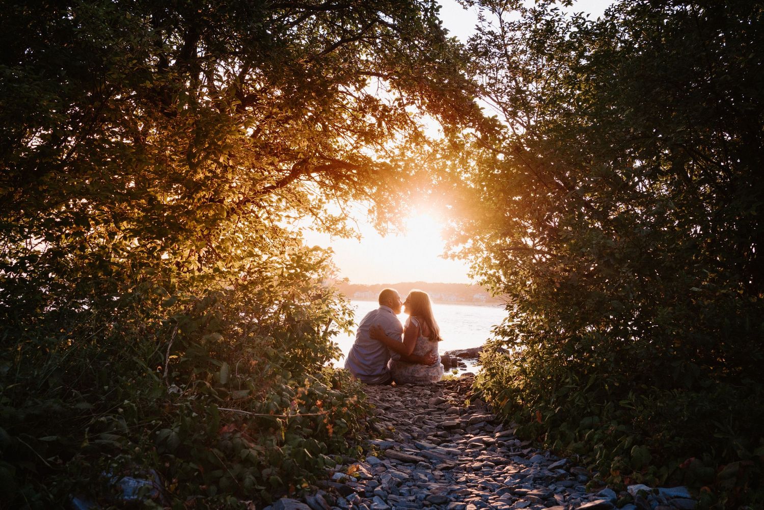 Silhouetted couple sits together on hilltop bench against dramatic golden sunset through trees.