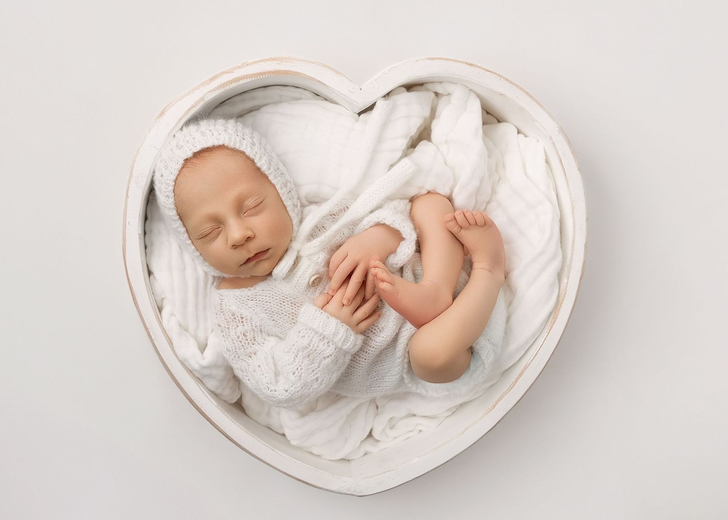 A sleeping newborn baby wrapped in soft white fabric lies curled in a round white bowl.