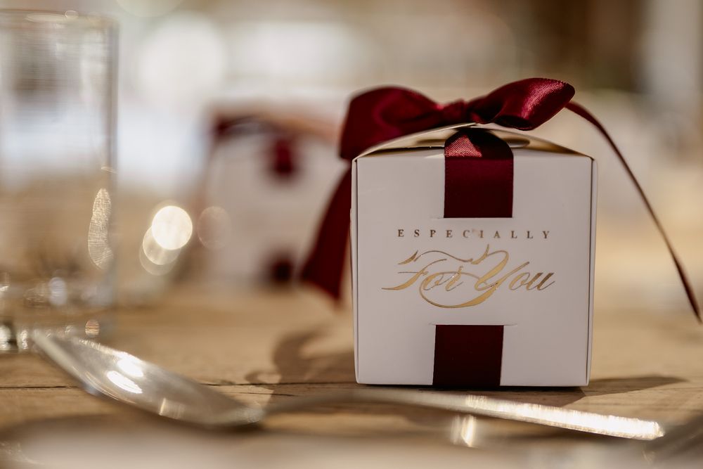 Elegant wedding favor box with burgundy ribbon and gold text sits on formal table setting.