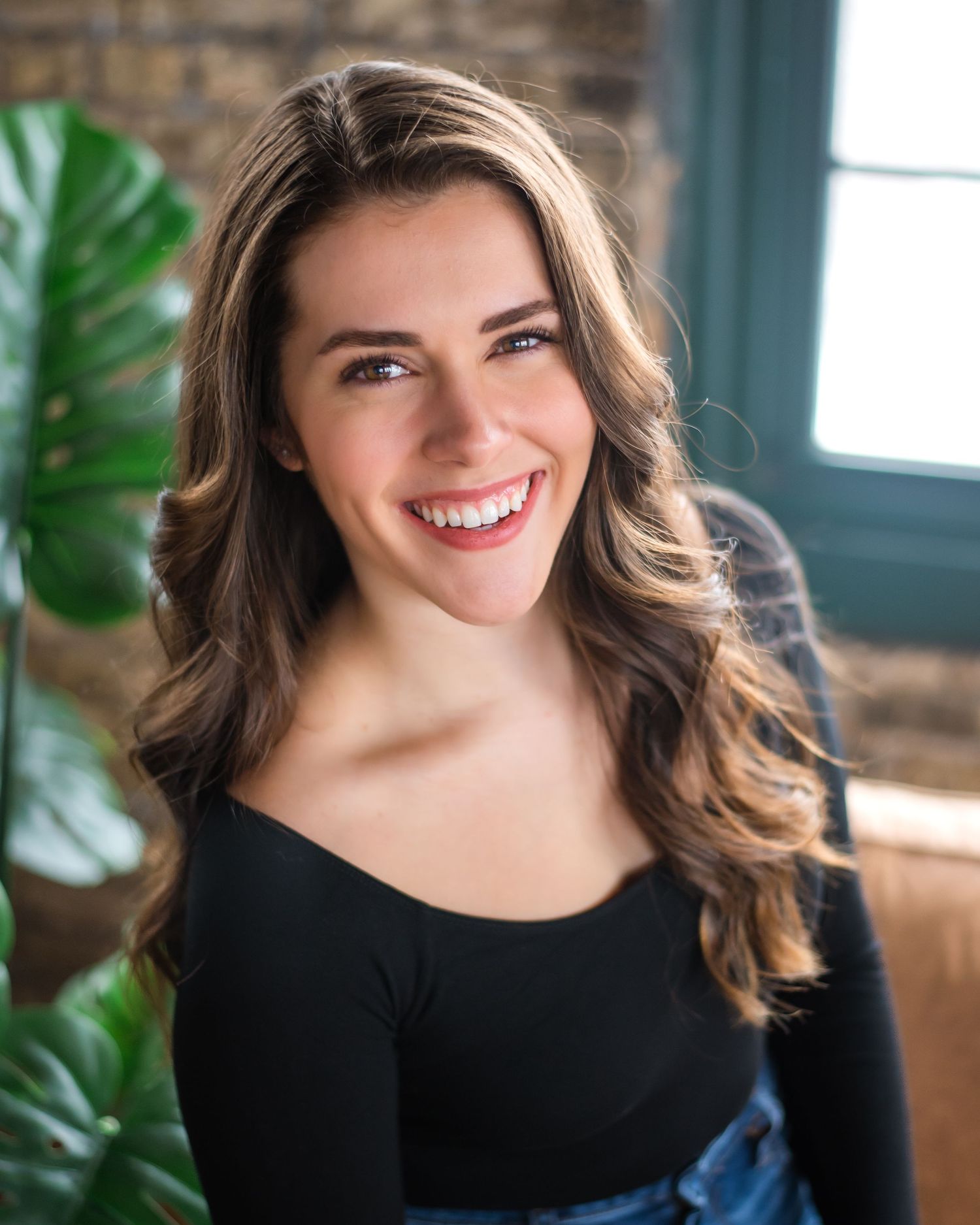 Professional portrait photo of someone with wavy brown hair and a bright smile wearing a black top in a sunlit room.
