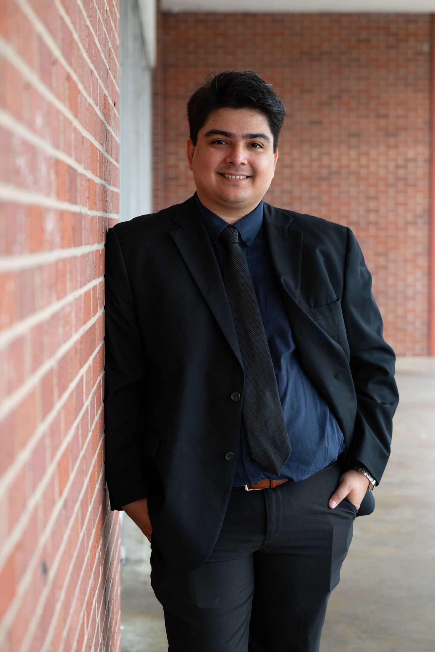 Professional portrait of a person in a dark suit and navy shirt standing against a brick wall.